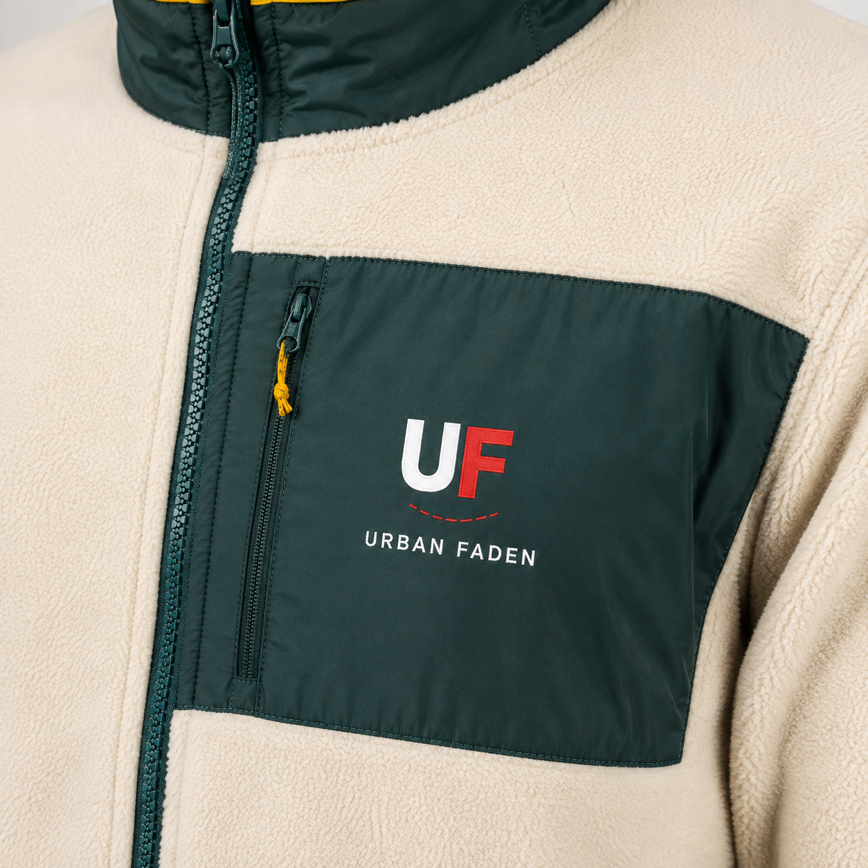 Nahaufnahme einer Fleecejacke mit dunkelgrüner Brusttasche und Reißverschluss, die ein weiß-rotes „UF“-Logo mit dem Schriftzug „URBAN FADEN“ auf der Tasche aufweist, und einem cremefarbenen Fleece-Körper.